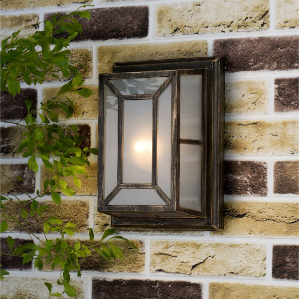 An outdoor wall light with a bronze finish and frosted glass panels, mounted on a brick wall with green foliage.