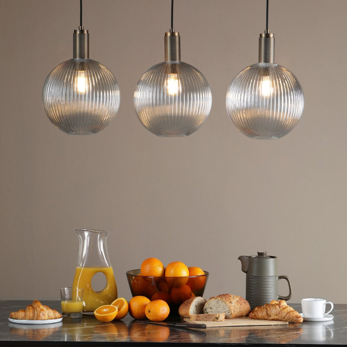 Three ribbed glass pendant lights illuminate a breakfast spread with croissants, oranges, juice, and bread.
