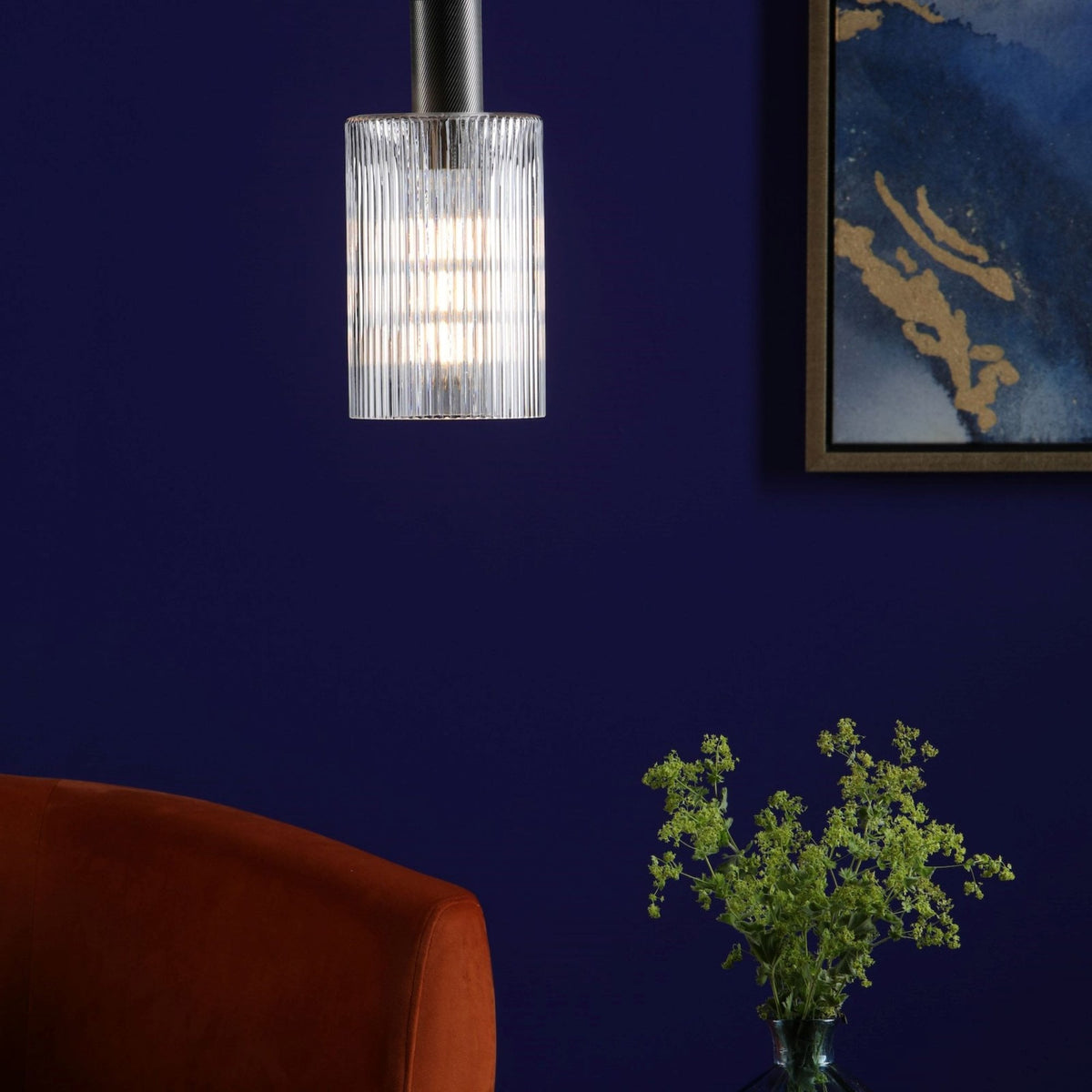 A fluted glass pendant light illuminates a dark blue wall above an orange chair and green plant.
