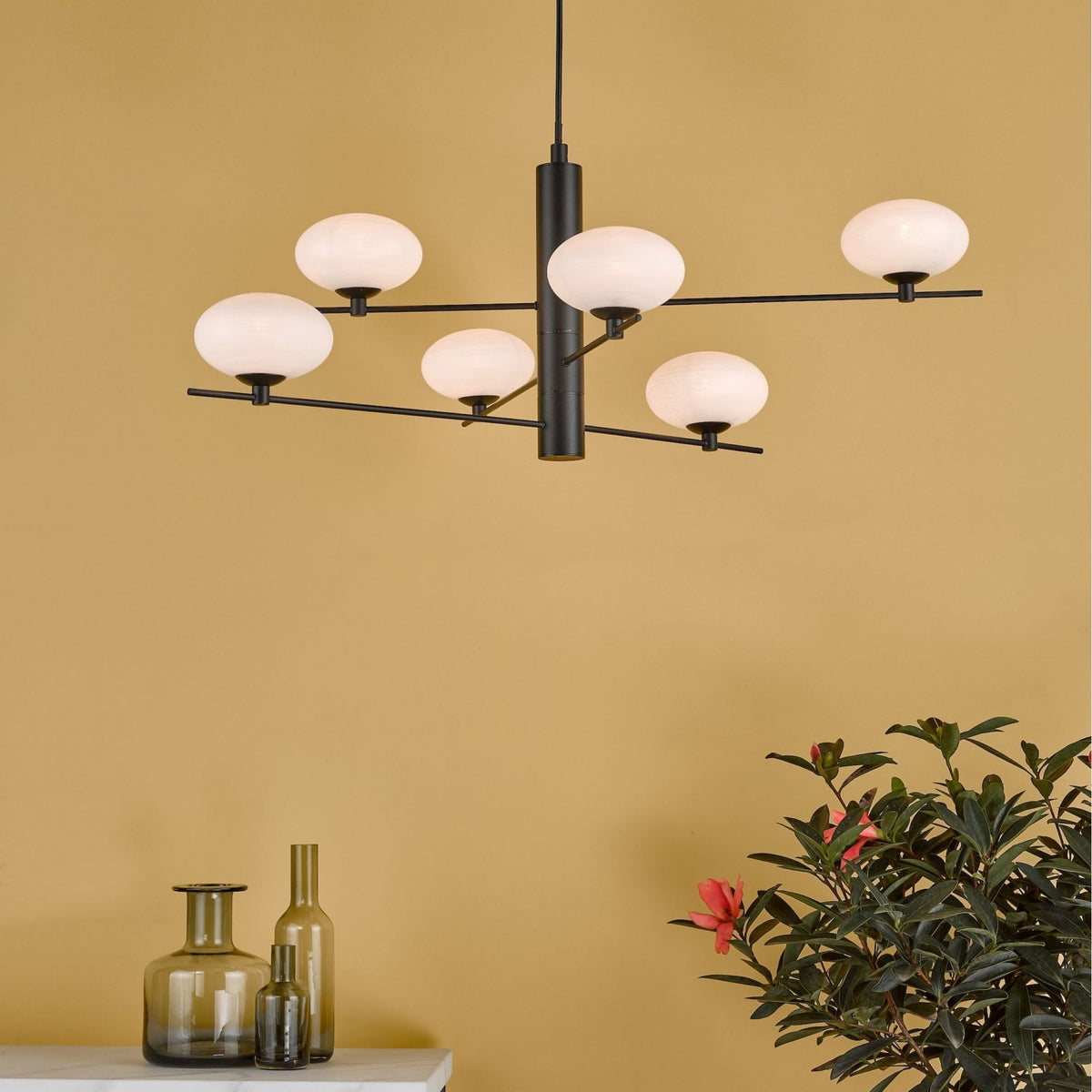 Modern black chandelier with six white globe lights, above green glass vases and a plant with pink flowers.