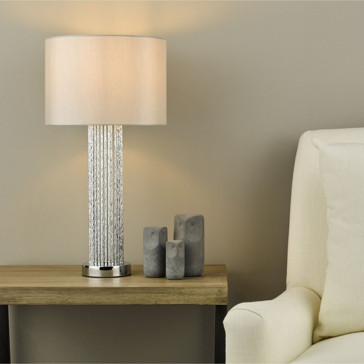 Table lamp with a textured silver base and a white shade, next to three owl figurines and a cream armchair.