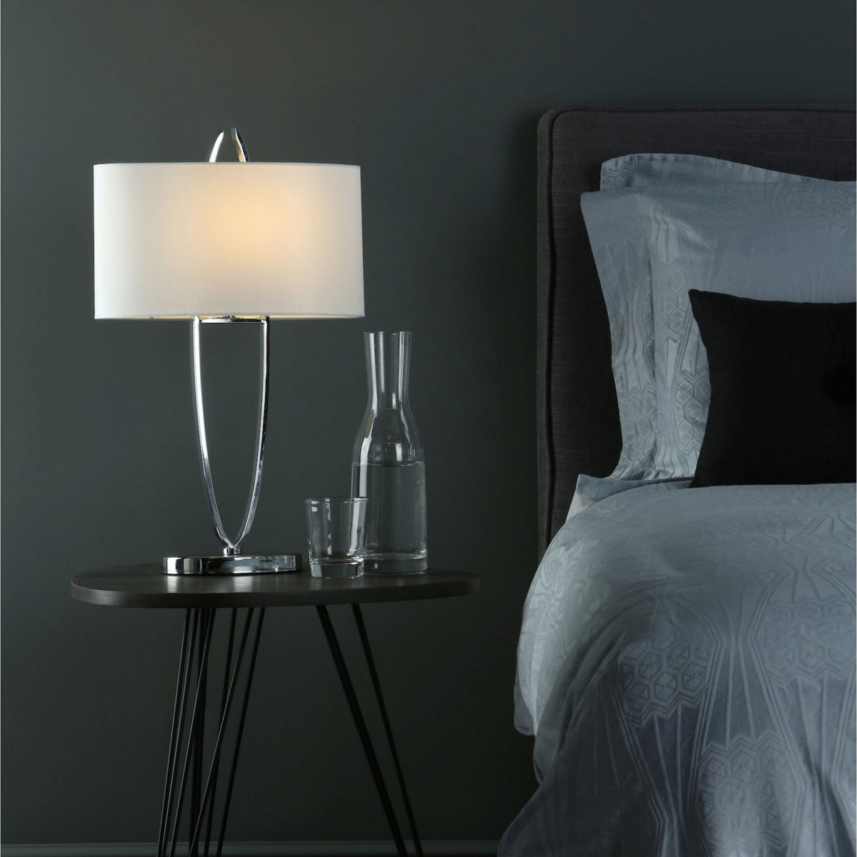 A modern bedroom with a chrome lamp, water carafe, and glass on a dark wood nightstand next to a bed with patterned bedding.