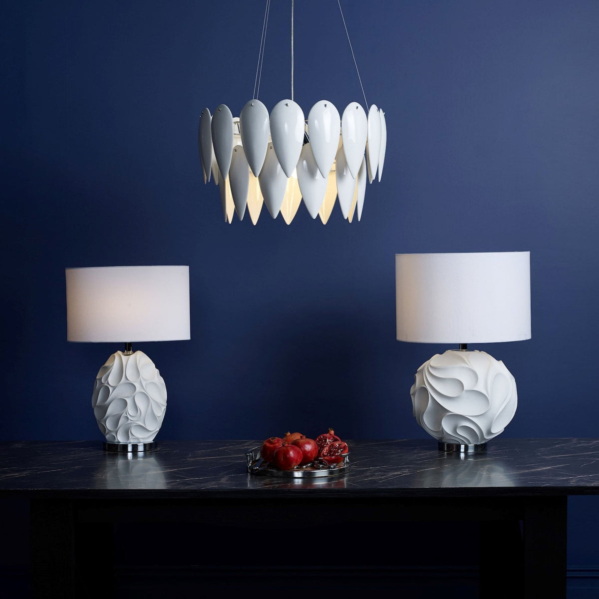 Two white table lamps with textured bases and a white chandelier with petal-like shades against a blue wall.
