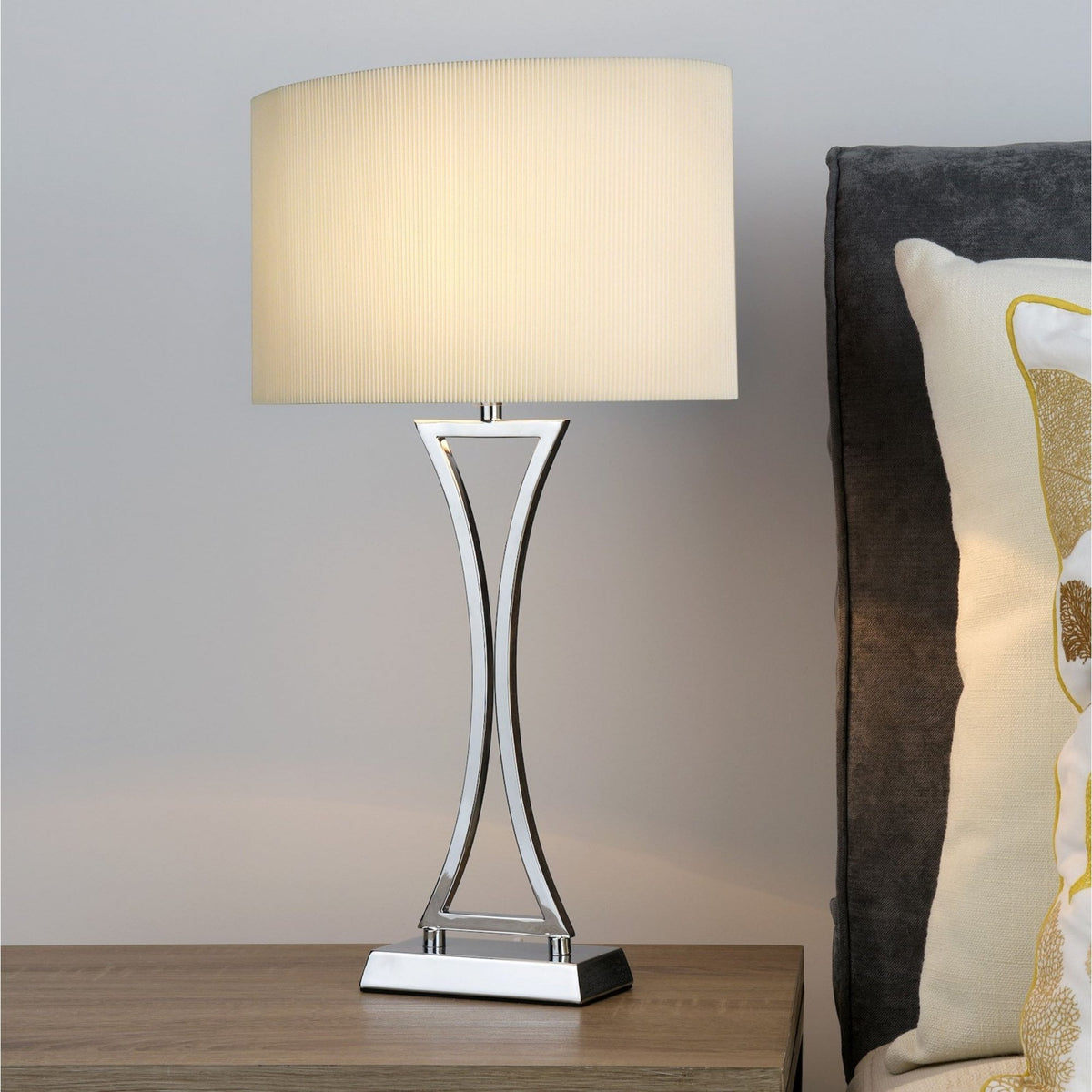 Modern table lamp with a chrome base and a rectangular pleated shade, casting a warm glow on a wooden nightstand.