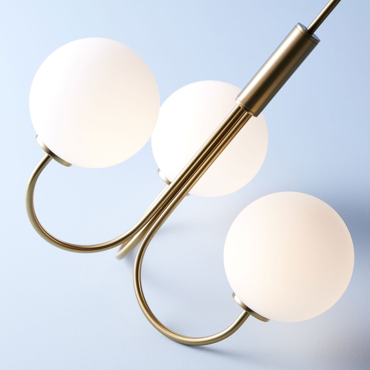 A modern brass chandelier with three glowing white globe shades on a pale blue background.