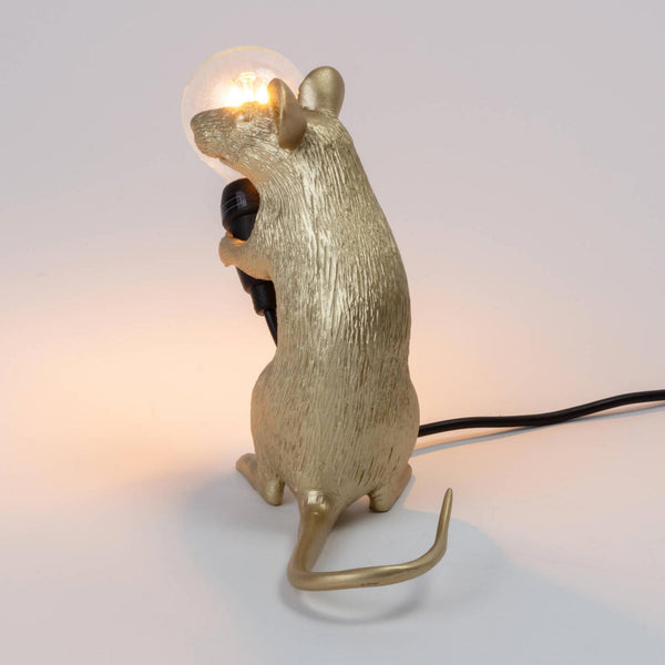 A golden mouse lamp holding a lit bulb, viewed from the back right side.