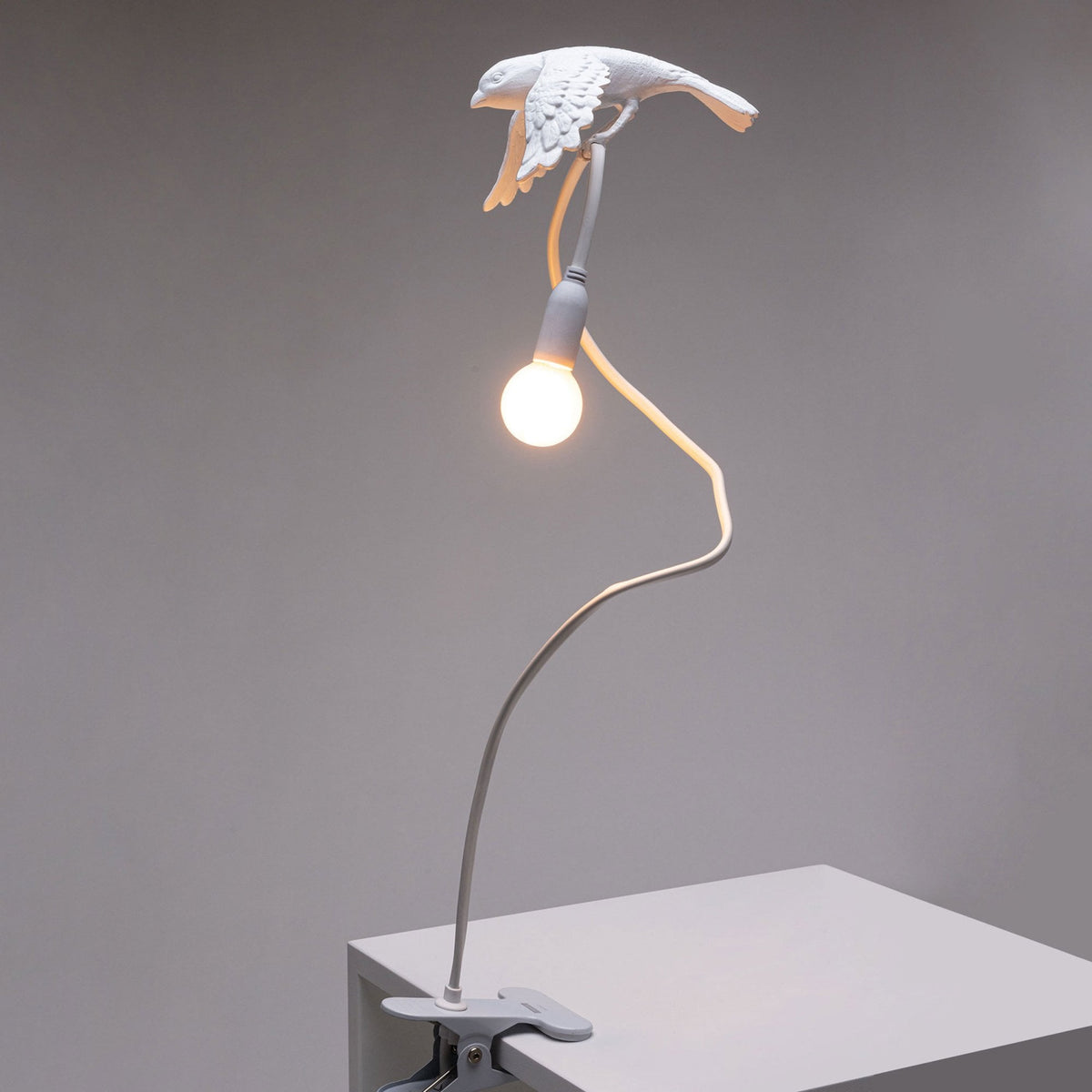 White bird lamp with illuminated bulb, perched on a long, flexible stand clipped to a surface.