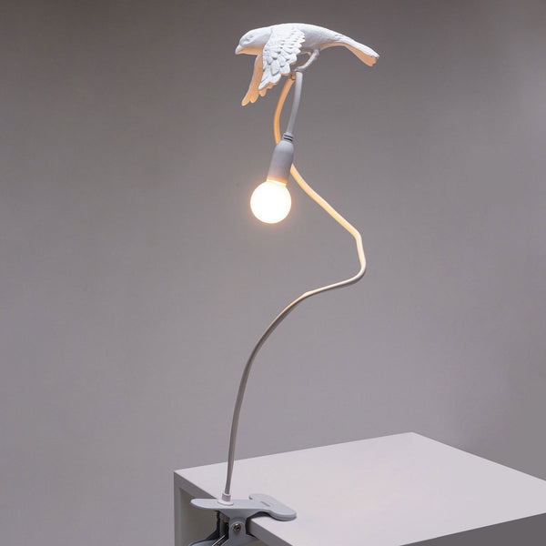 White bird lamp with illuminated bulb, perched on a long, flexible stand clipped to a surface.