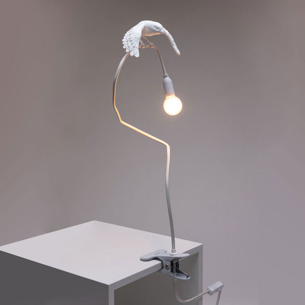 Elegant white bird lamp with a glowing bulb, on a flexible arm, clipped to a table edge.