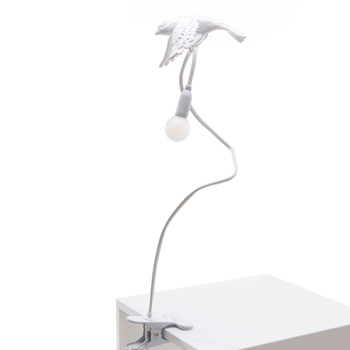White bird lamp with illuminated bulb on a flexible stand, clipped to the edge of a white surface.