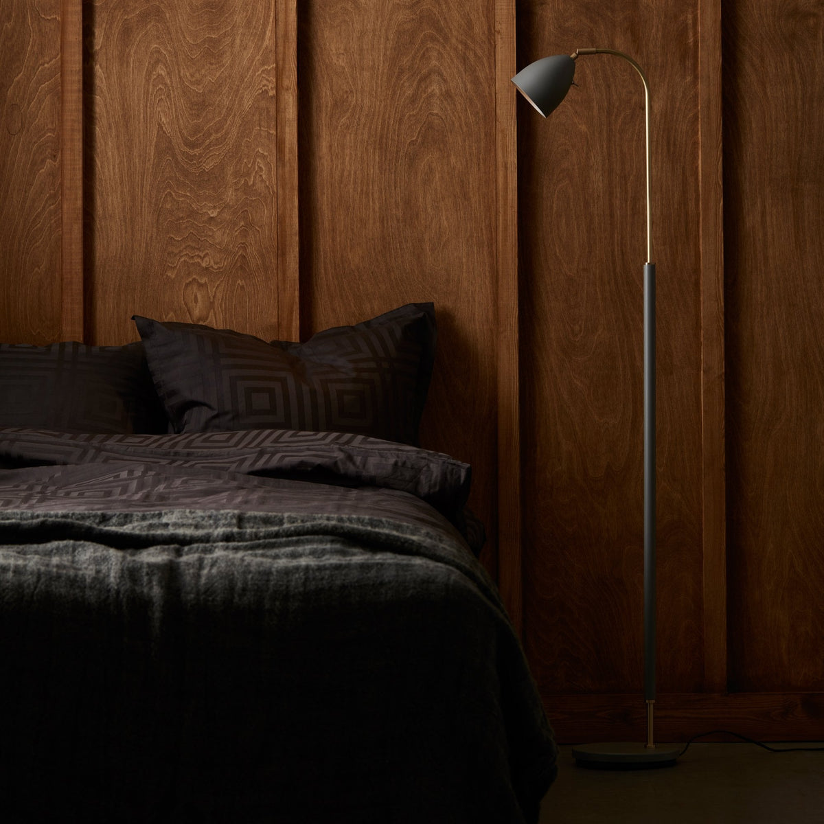 BELID Deluxe Floor Lamp in Warm Grey & Brass next to a bed with dark bedding and a wooden wall