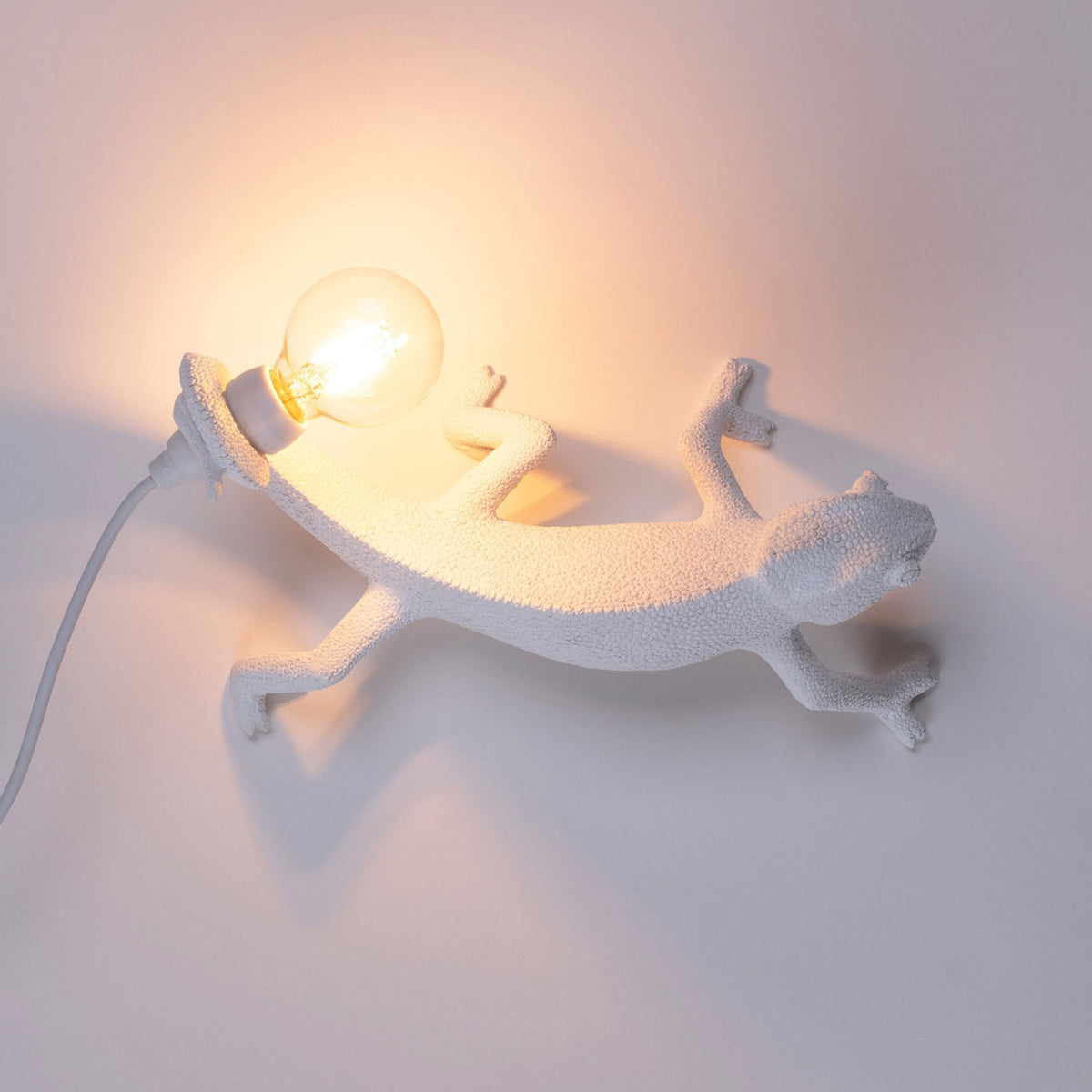 A white chameleon-shaped wall lamp with an exposed bulb illuminates a plain white wall.