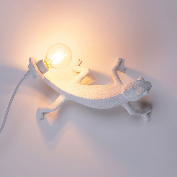 A white chameleon-shaped wall lamp with an exposed bulb illuminates a plain white wall.