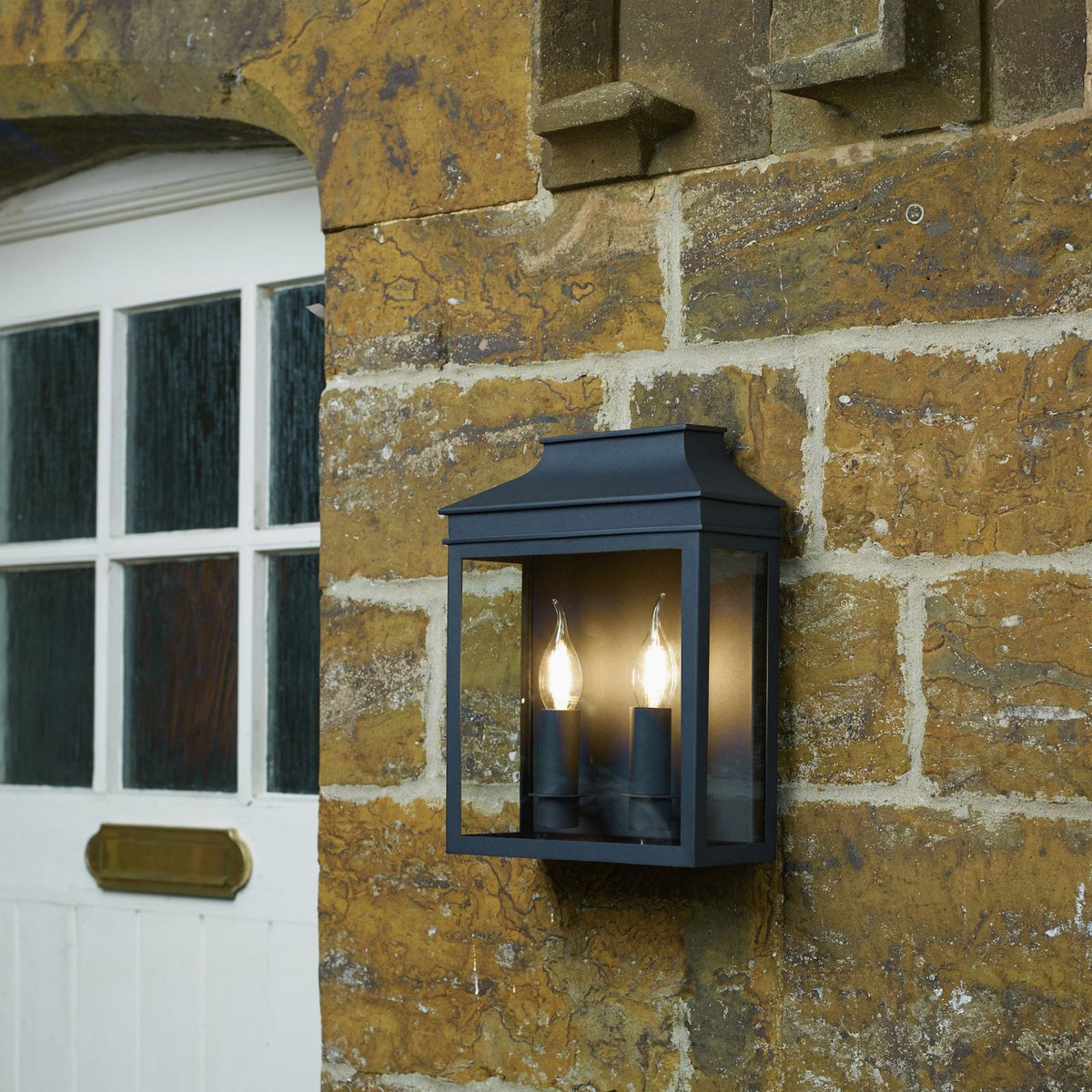 A black outdoor lantern with two glowing bulbs mounted on a rustic stone wall next to a white door.