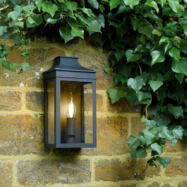 A black outdoor wall light with clear glass panels, installed on a stone wall amidst lush ivy.