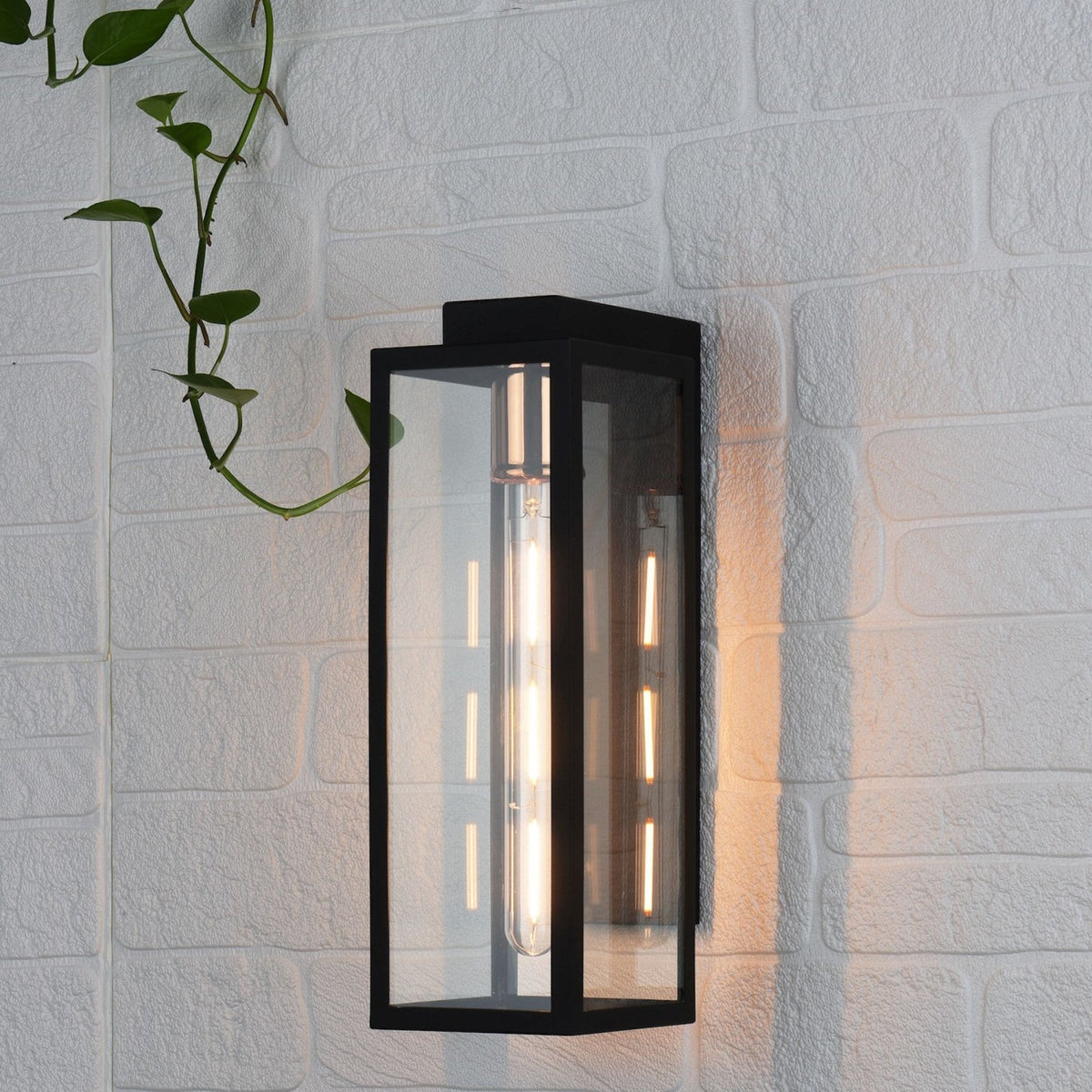 Black rectangular wall sconce with a clear glass shade and exposed filament bulb on a white brick wall.