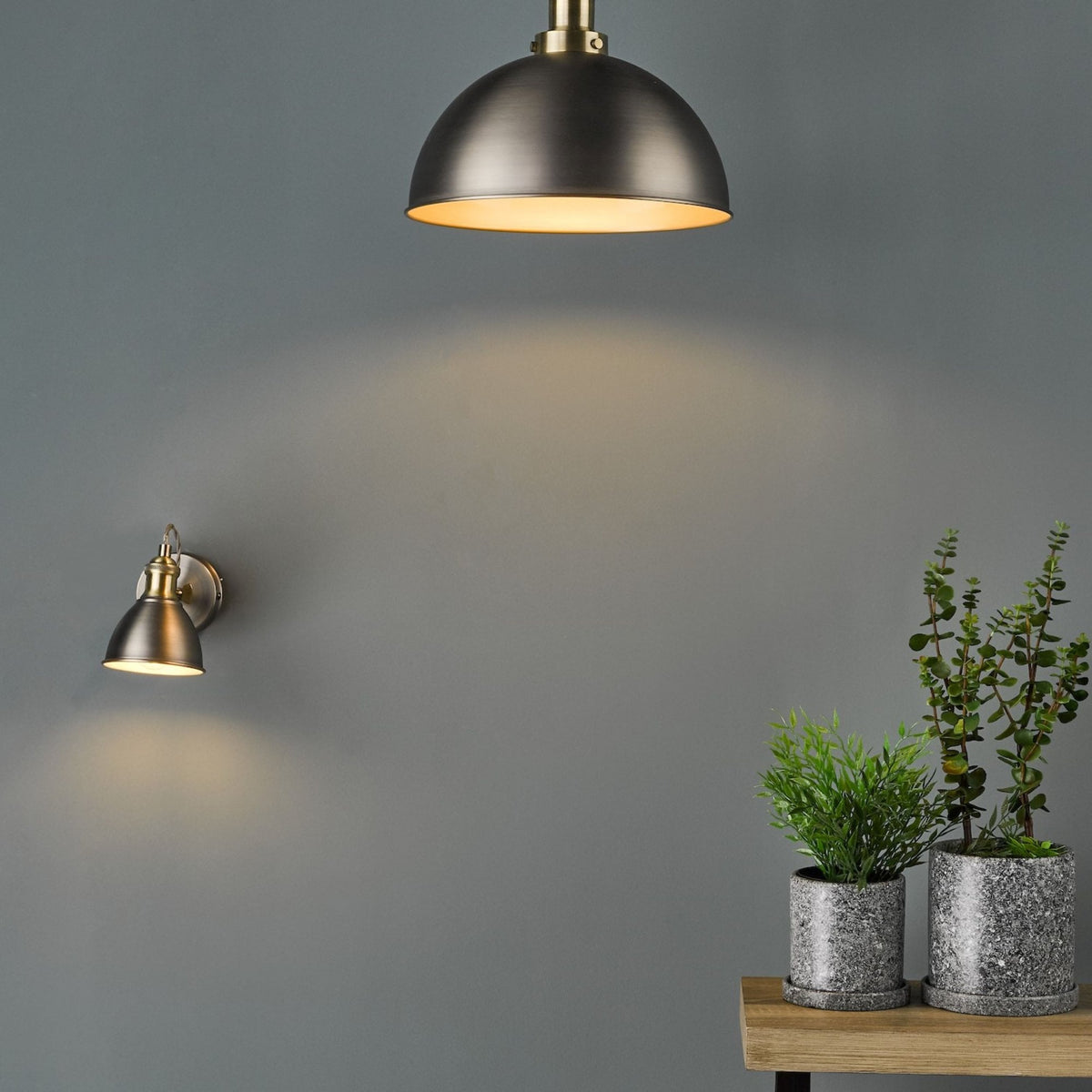 Two antique brass lights illuminate a gray wall above potted plants on a wooden shelf.