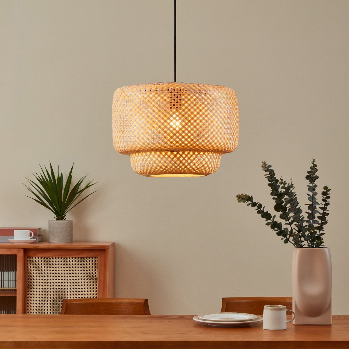 A bamboo pendant light illuminates a dining room with a wooden table and plants.