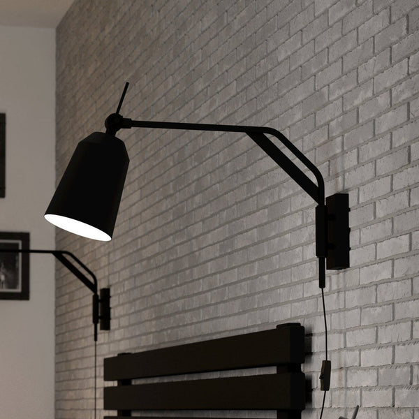 A black wall lamp with an adjustable arm and conical shade illuminates a grey brick wall.