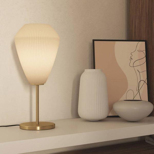 A white pleated table lamp with a brass base and stem sits on a shelf with decorative items.