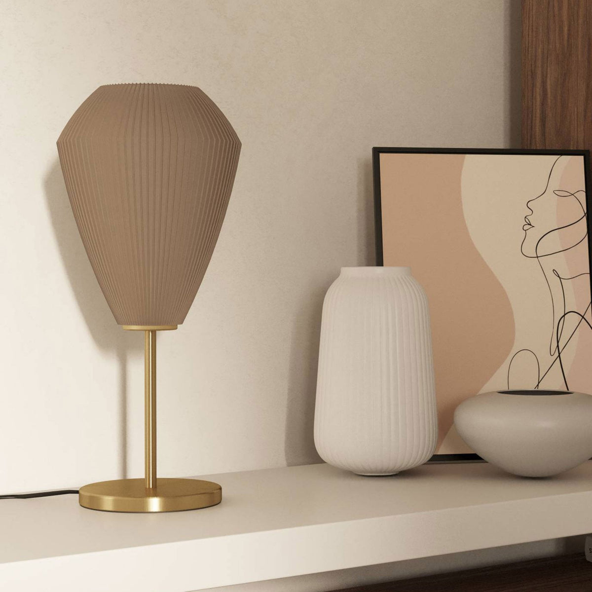 A beige pleated table lamp with a brass base and stem on a shelf next to decorative items.