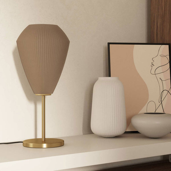 A beige pleated table lamp with a brass base and stem on a shelf next to decorative items.