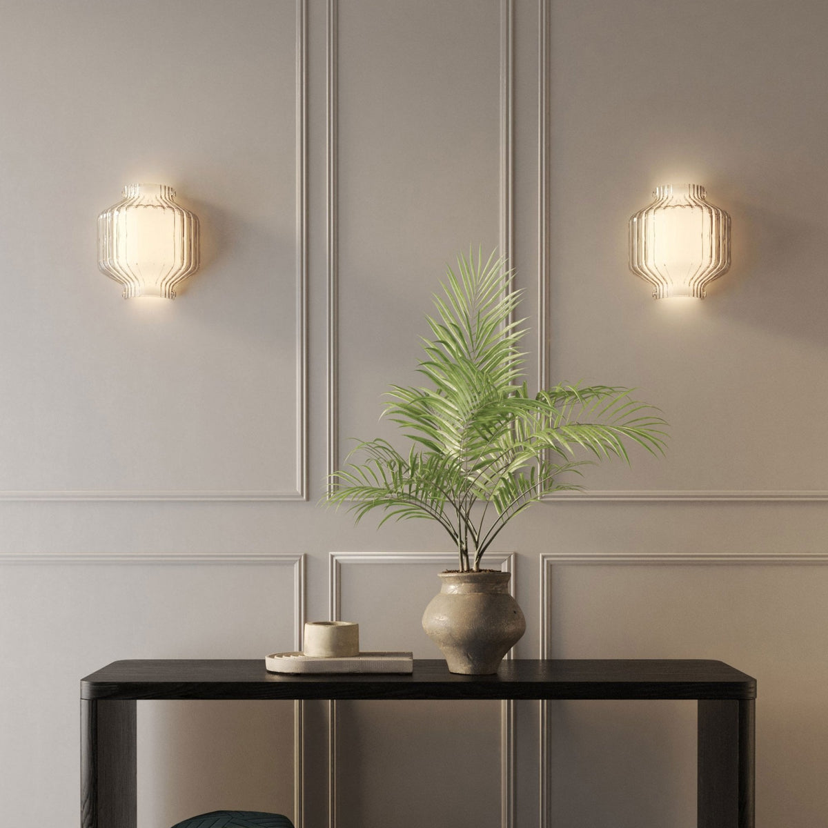 Two illuminated glass wall sconces flank a potted plant on a dark console table against a paneled wall