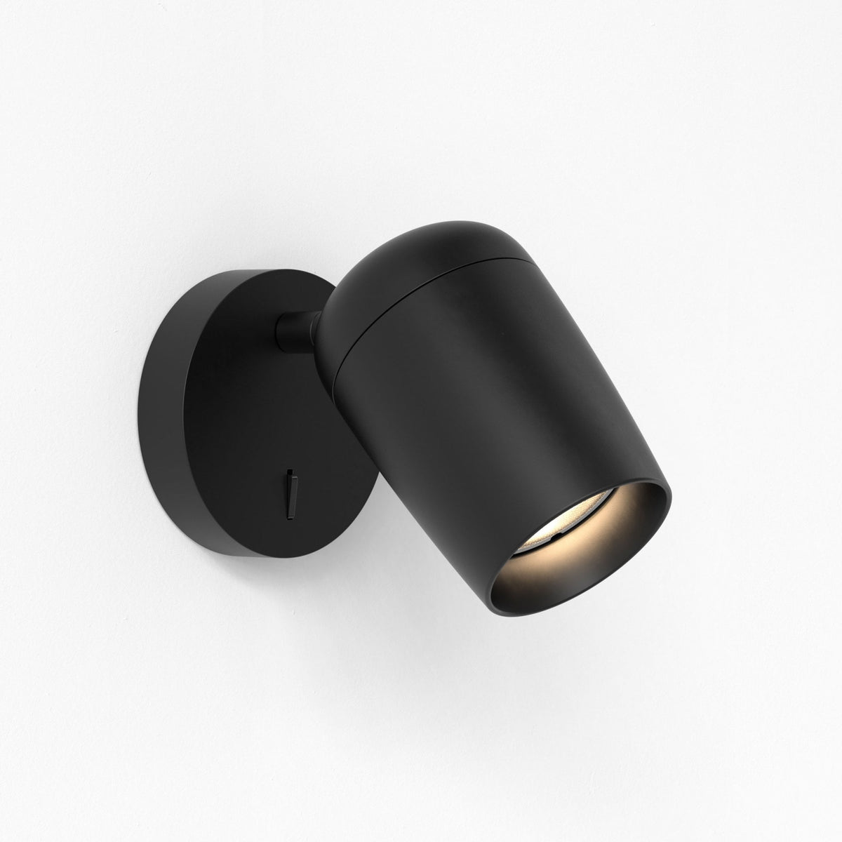 Black wall-mounted spotlight with a cylindrical head angled downwards, emitting a warm light