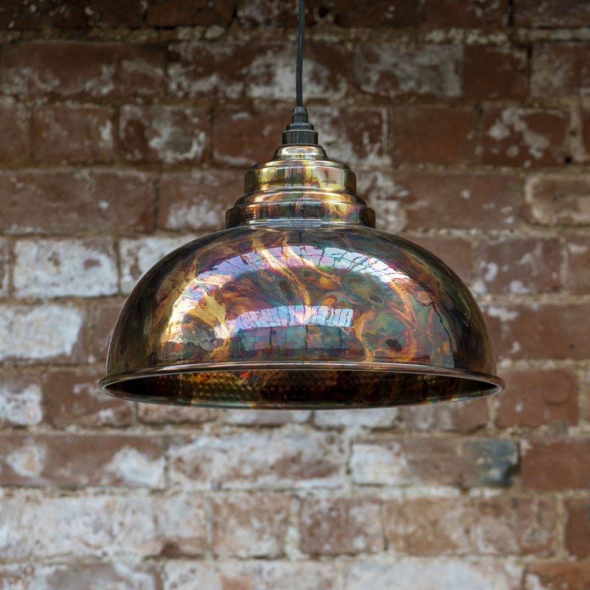 Dome pendant light with an iridescent, multicoloured, metallic shade against a brick wall.