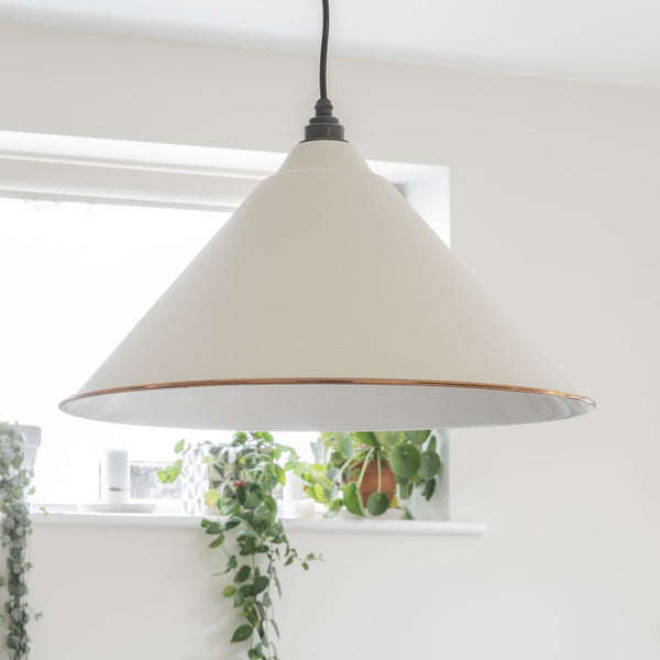 A white conical pendant light with a copper band hangs in front of a window with plants.
