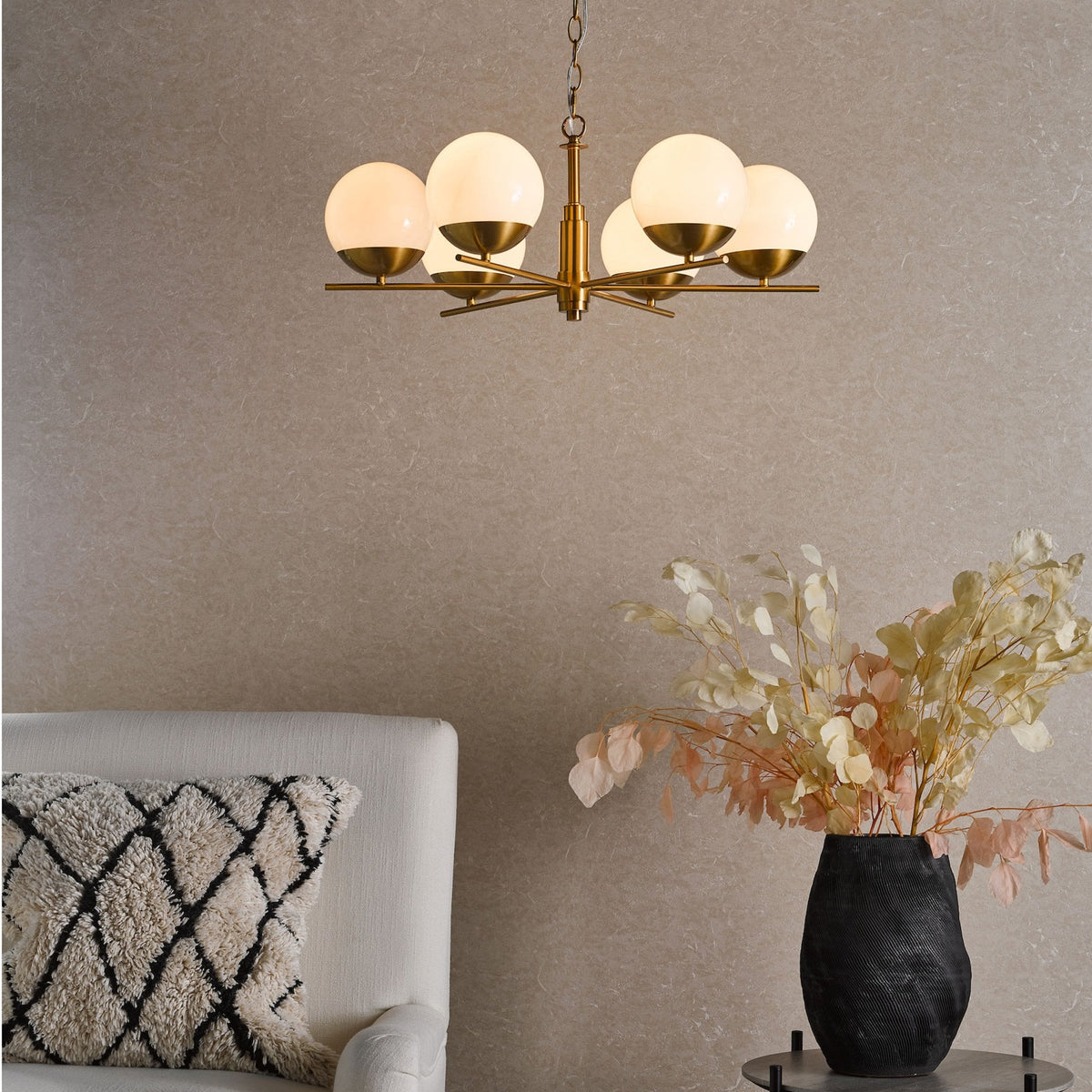 A modern brass chandelier with five globe lights illuminates a room with a textured wall, a white armchair, and a black vase with dried flowers.