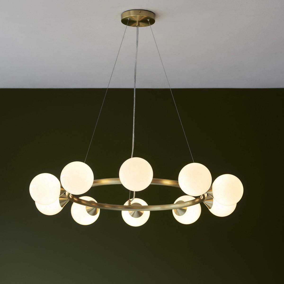 A round brass chandelier with ten white globe lights hangs from a ceiling against a dark green background.