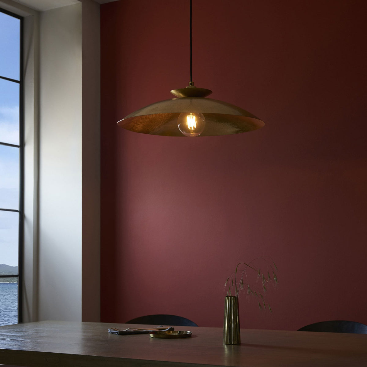 A gold-colored pendant lamp hanging above a wooden table with a brass vase, against a red wall and window.