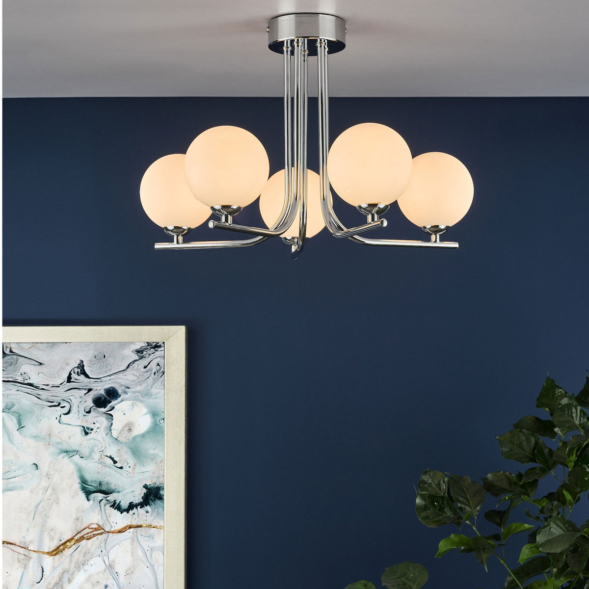 A modern chrome ceiling light with five spherical white glass shades illuminates a dark blue wall.