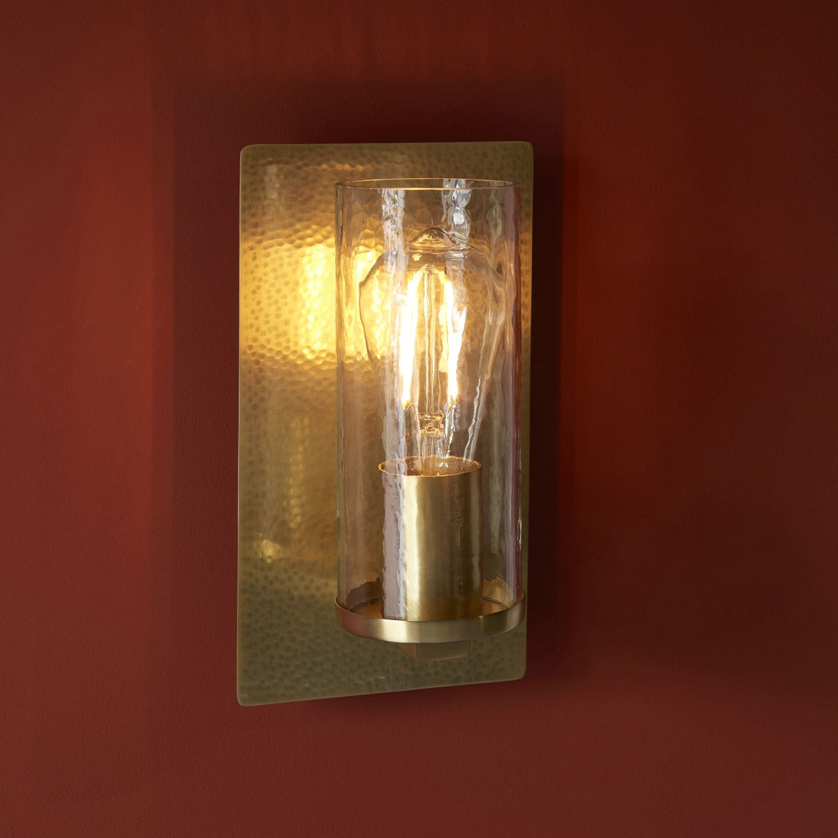 A close up view of a rectangular, hammered gold wall sconce with a clear glass cylinder and an exposed bulb.