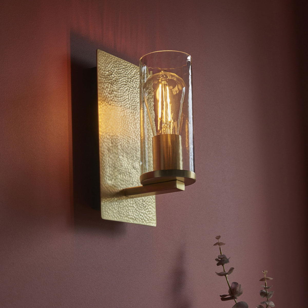 A rectangular hammered gold wall sconce with a clear glass cylinder and exposed bulb lights a dark plum wall.