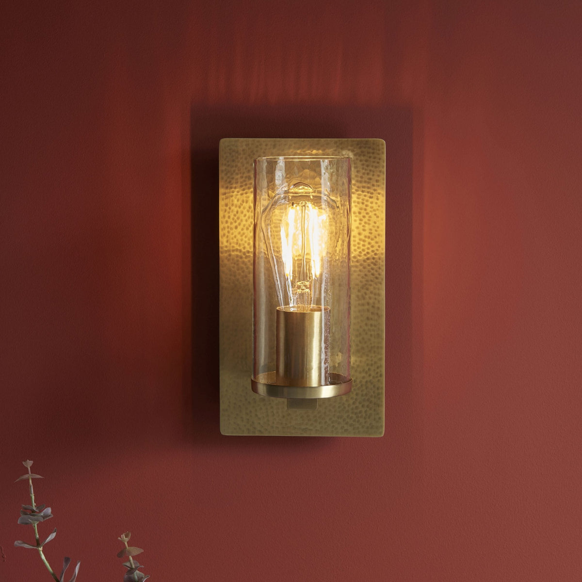 A rectangular, hammered gold wall sconce with a clear glass cylinder and exposed bulb lights up a red wall.