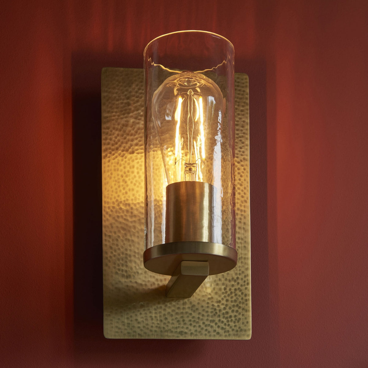 A rectangular, hammered gold wall sconce with a clear glass cylinder and exposed bulb against a red wall.