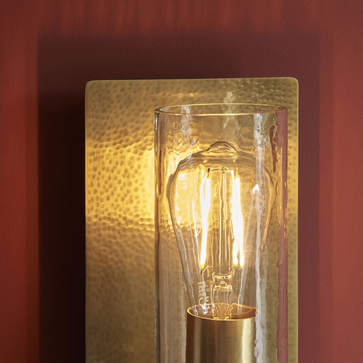 A close up view of a rectangular, hammered gold wall sconce with a clear glass cylinder and exposed bulb.