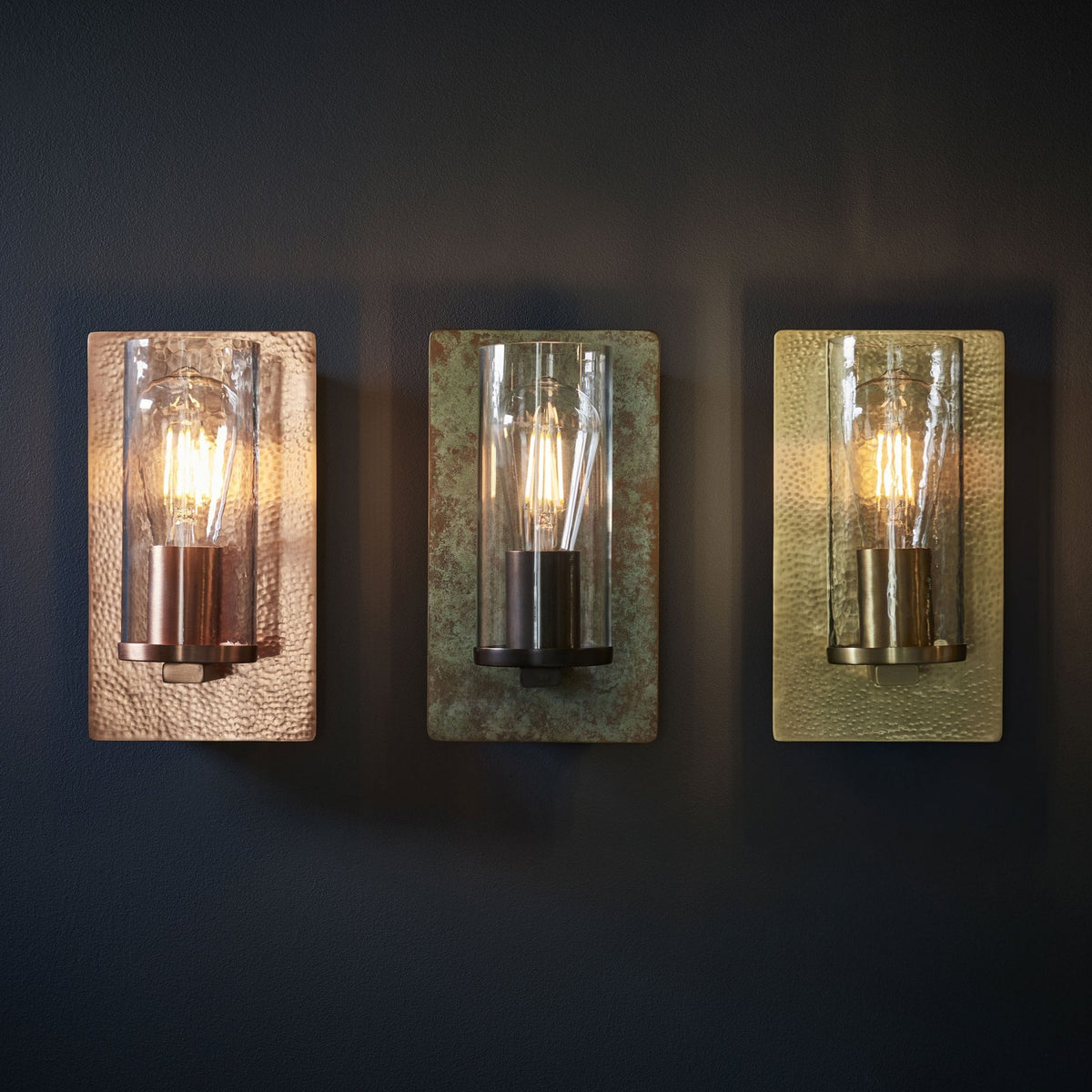 Three rectangular wall sconces with clear glass cylinders and exposed bulbs are displayed against a dark wall.