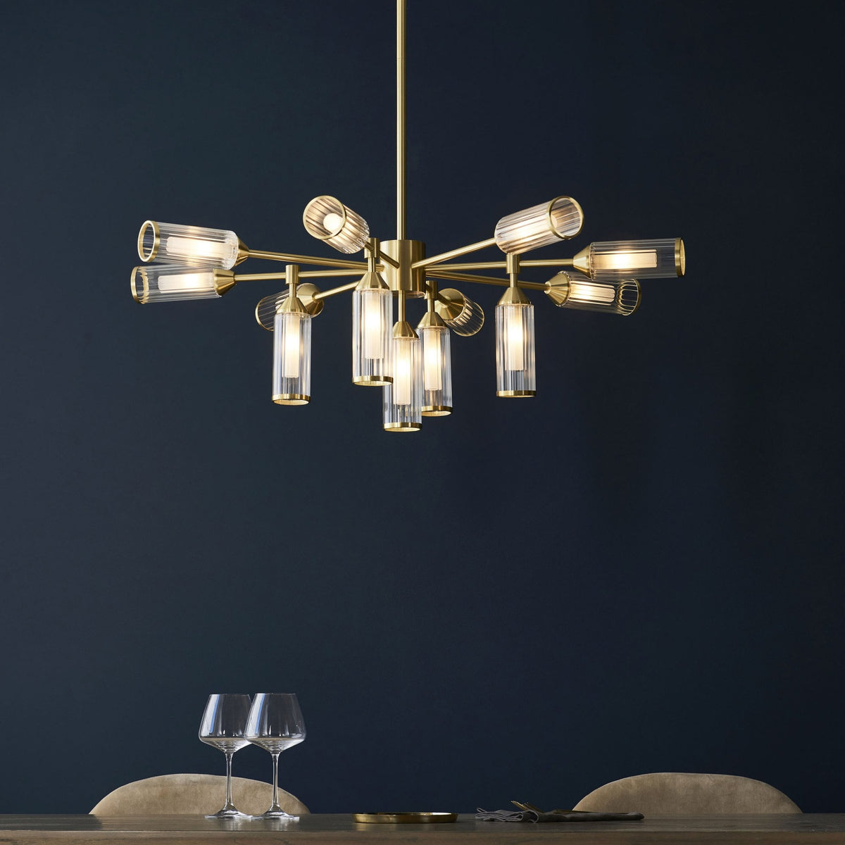 A sleek gold chandelier with clear and ribbed glass shades illuminates a dining area with wine glasses.