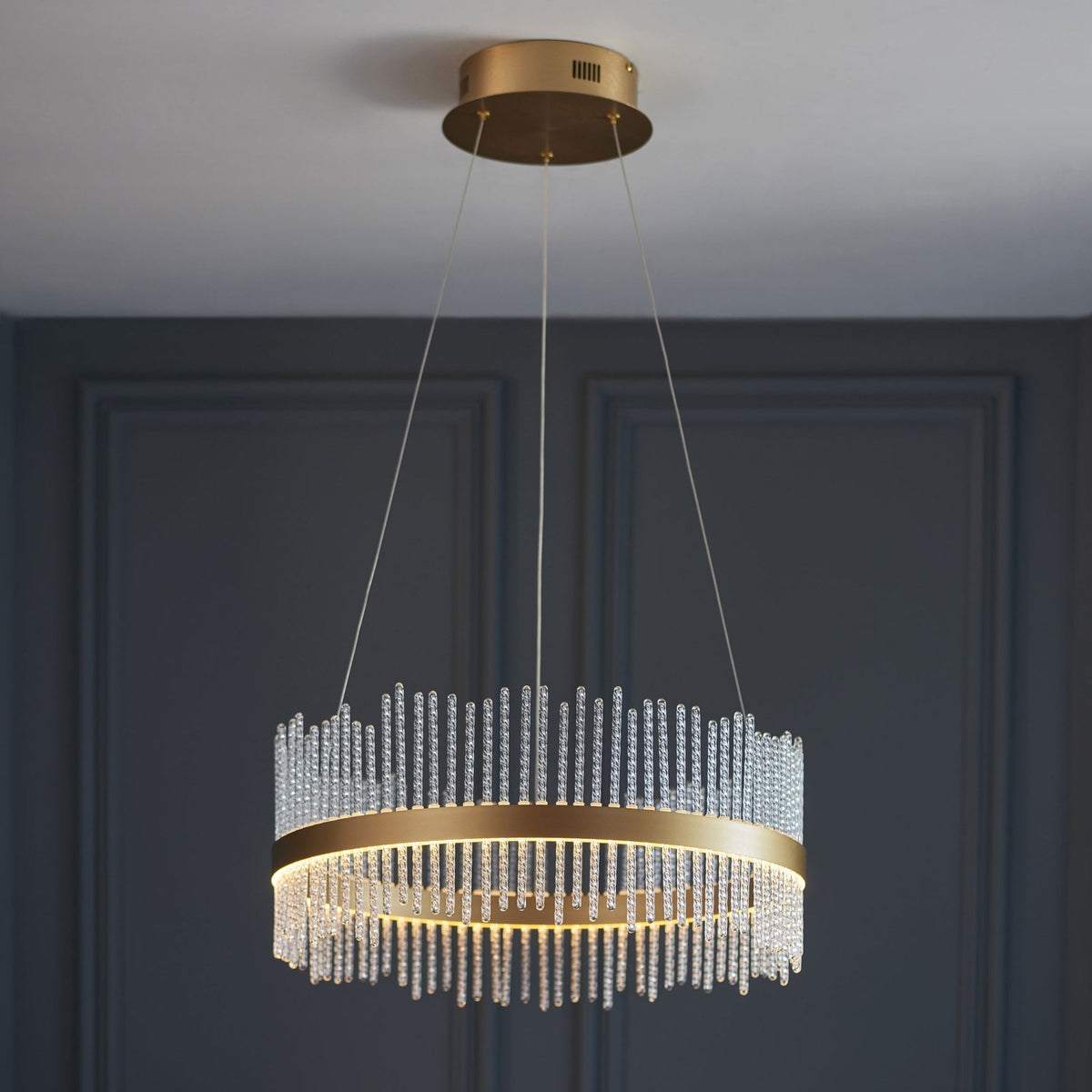 A round, brass-finished chandelier with multiple clear glass rods hanging from it, set against a dark wall.