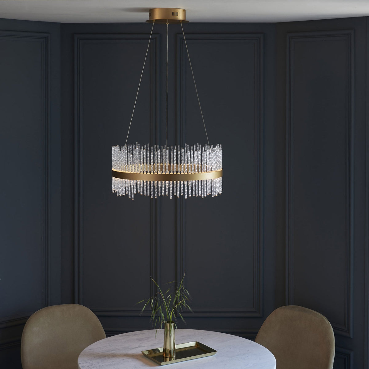 A golden chandelier with hanging glass rods illuminating a dining area with a round table and two light-colored chairs.