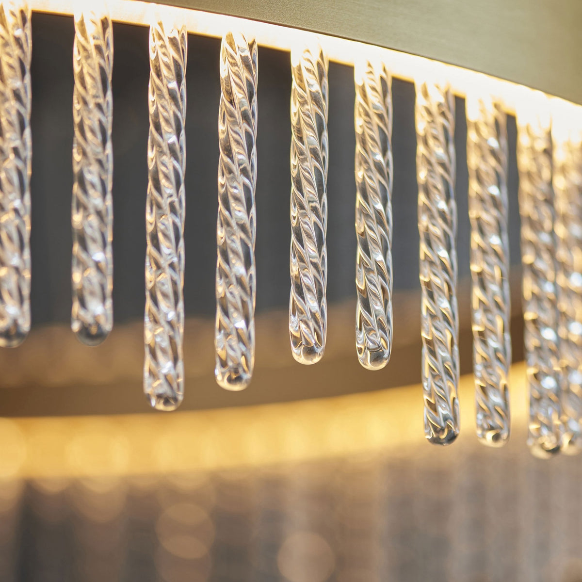 A close-up of clear glass rods with internal textures hanging from a golden chandelier, illuminated from above.