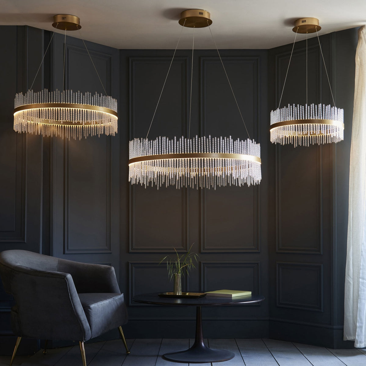 Three brass chandeliers with glass rods, two round and one rectangular, illuminate a modern living space.