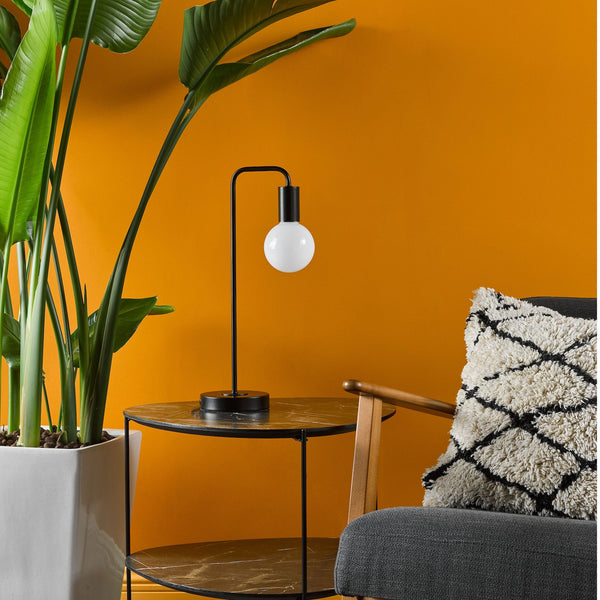 Modern black table lamp with a white bulb on a two-tier marble side table next to a large plant.