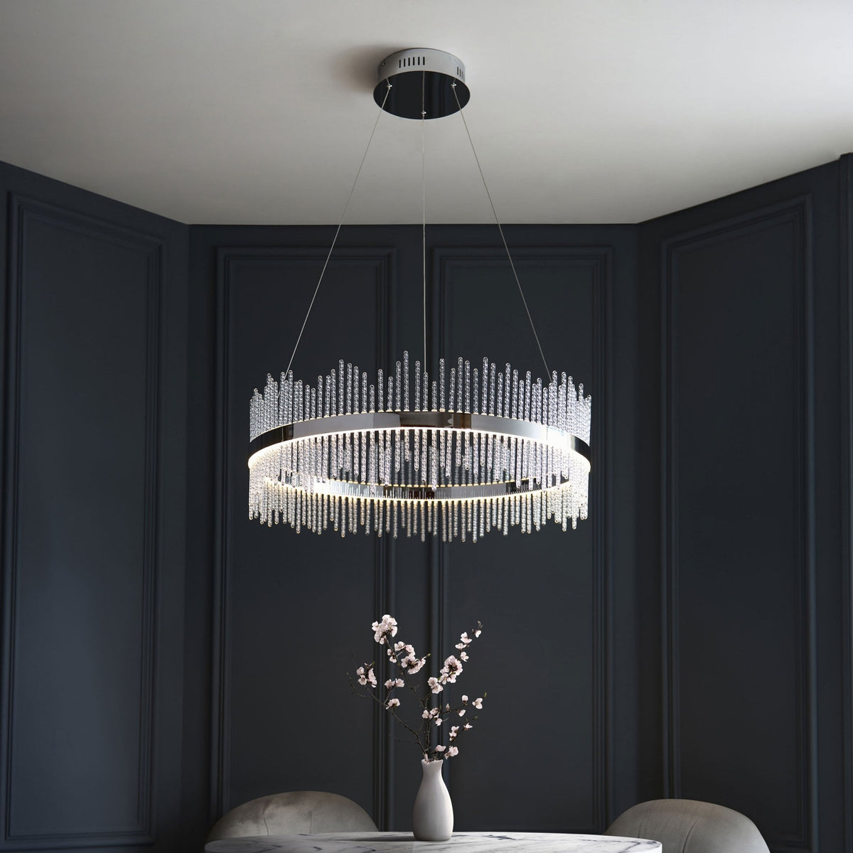 A luxurious crystal chandelier hanging above a dining table with a vase of flowers, illuminating a dark room.