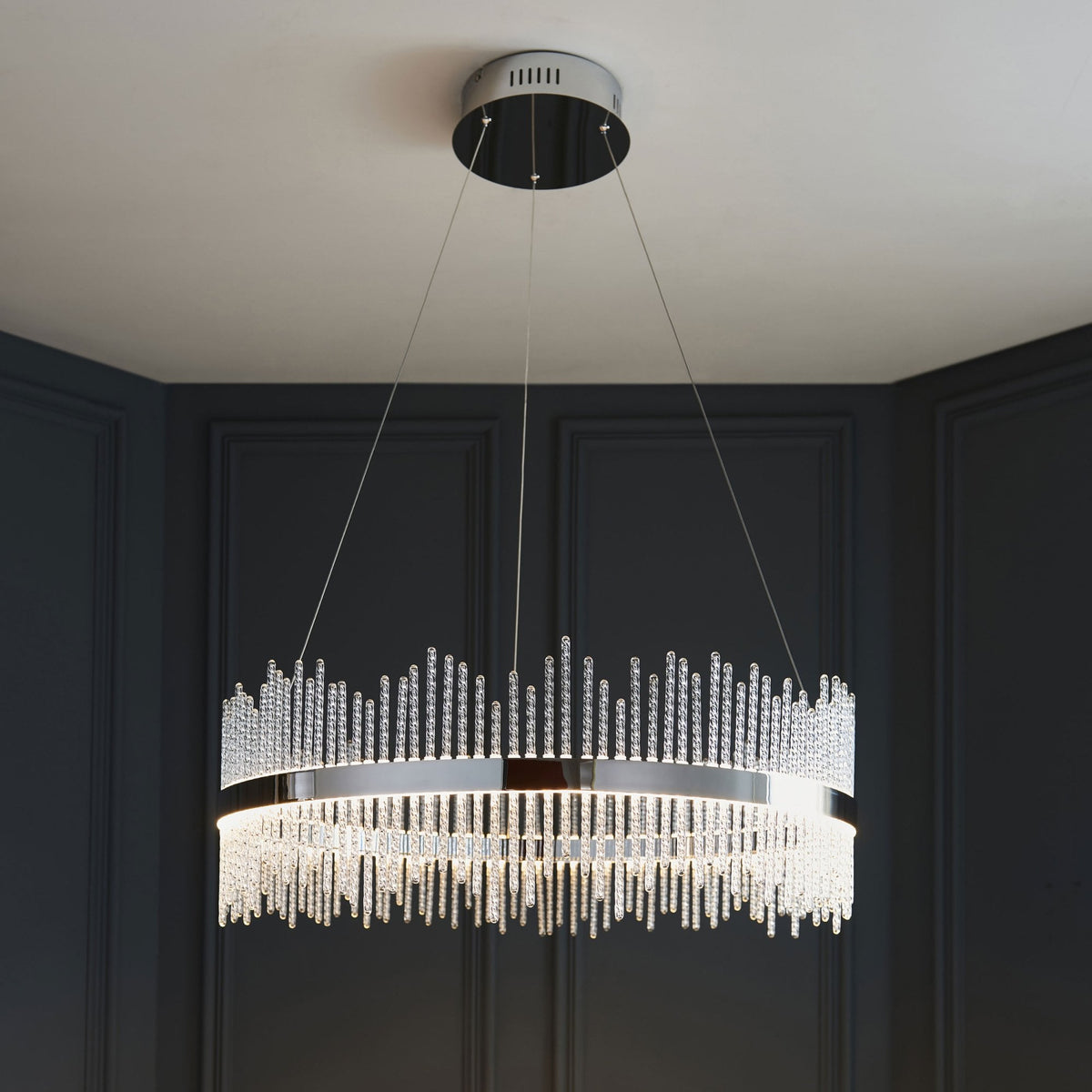 A modern crystal chandelier illuminating a dining area with dark paneled walls and a table with flowers.