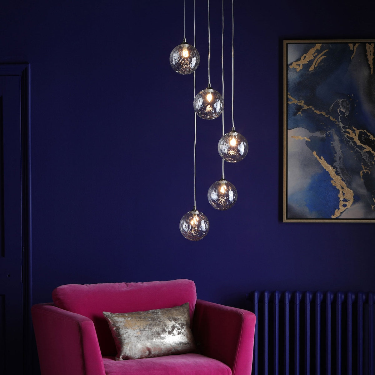 Five glass orb pendant lights hang over a pink velvet armchair with a gold cushion.