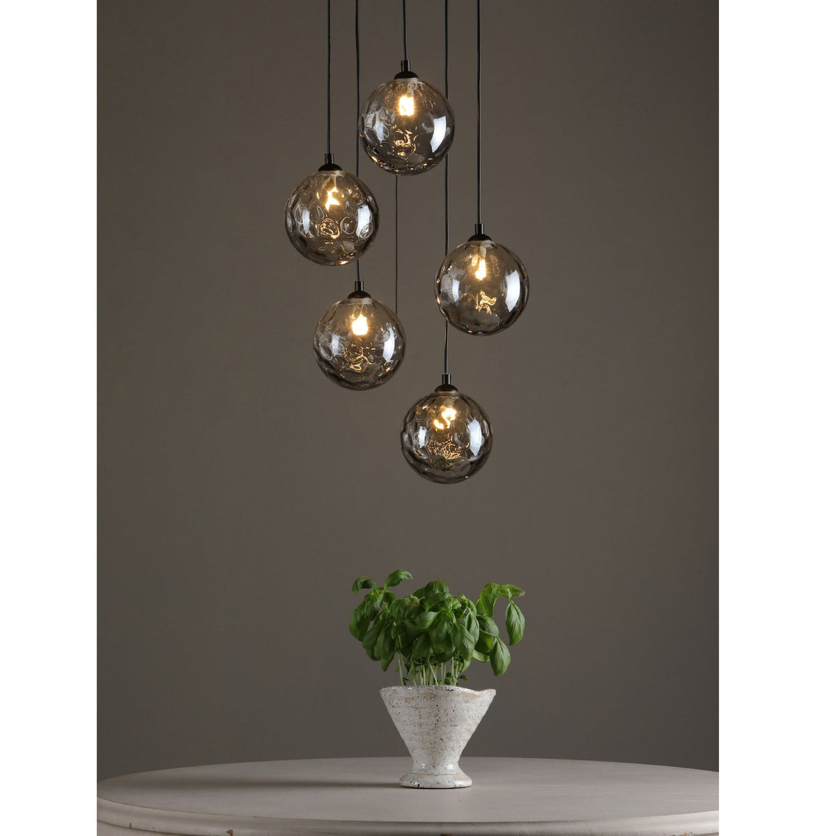 Five textured glass pendant lights hang above a potted basil plant on a round table.